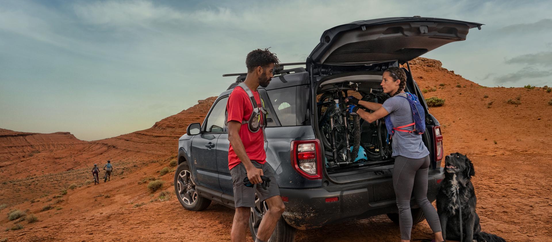 Two hikers loading mountain bikes into a Ford Bronco Sport beside a black dog
